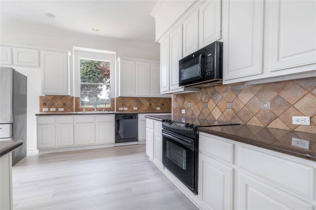 This is a bright, modern kitchen with white cabinetry, stainless steel appliances, and a tawny brown tiled backsplash. It features a window above the sink letting in natural light and has a sleek, gray-toned laminate flooring.