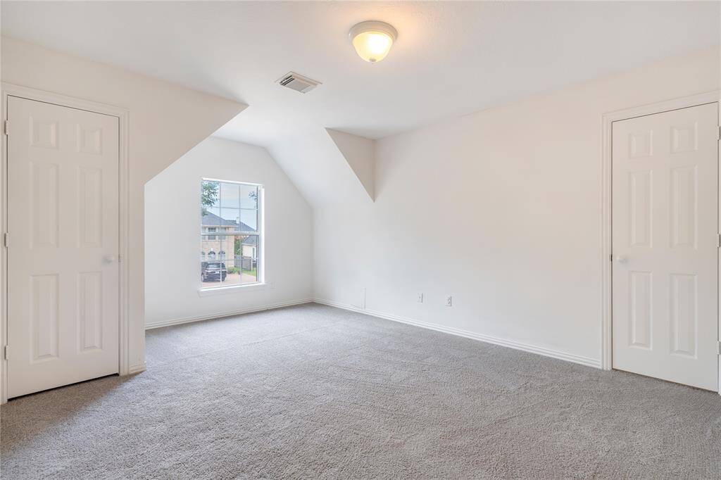 This is a spacious, well-lit room with neutral carpeting, featuring a large window for natural light, and it has two doors, possibly leading to a closet and an en suite bathroom. The angled ceiling adds architectural interest.
