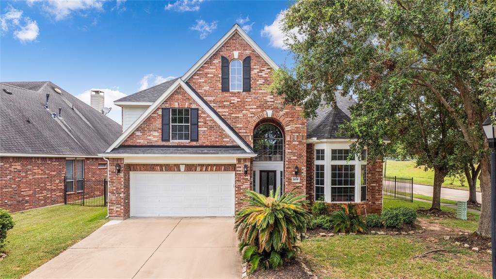 This is a two-story brick home  featuring a pitched roof with a prominent gable, arched entryway, and a two-car garage. The house features large windows, a well-maintained lawn, and mature trees, providing shade and curb appeal.