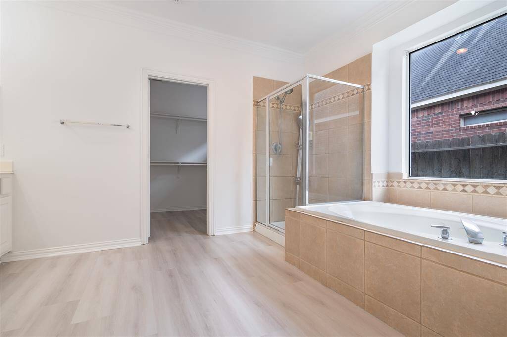 This is a bright, clean bathroom featuring a large built-in bathtub with shower, beige tile surround, and a window for natural light. There's also a built-in closet for storage. The flooring is a light, wood-look laminate.
