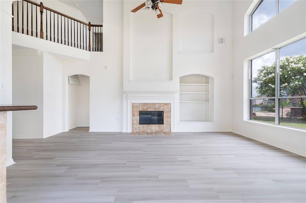 This is a spacious living room with high ceilings, ample natural light from large windows, a fireplace, and built-in shelving, featuring modern light wood flooring and a view of the upper-level walkway.