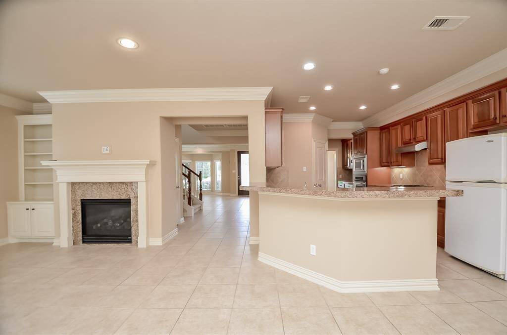 This spacious open-plan living area features a modern kitchen with wood cabinetry and a large center island. Adjacent is a cozy living space with a fireplace and built-in shelving, all set on elegant tile flooring.
