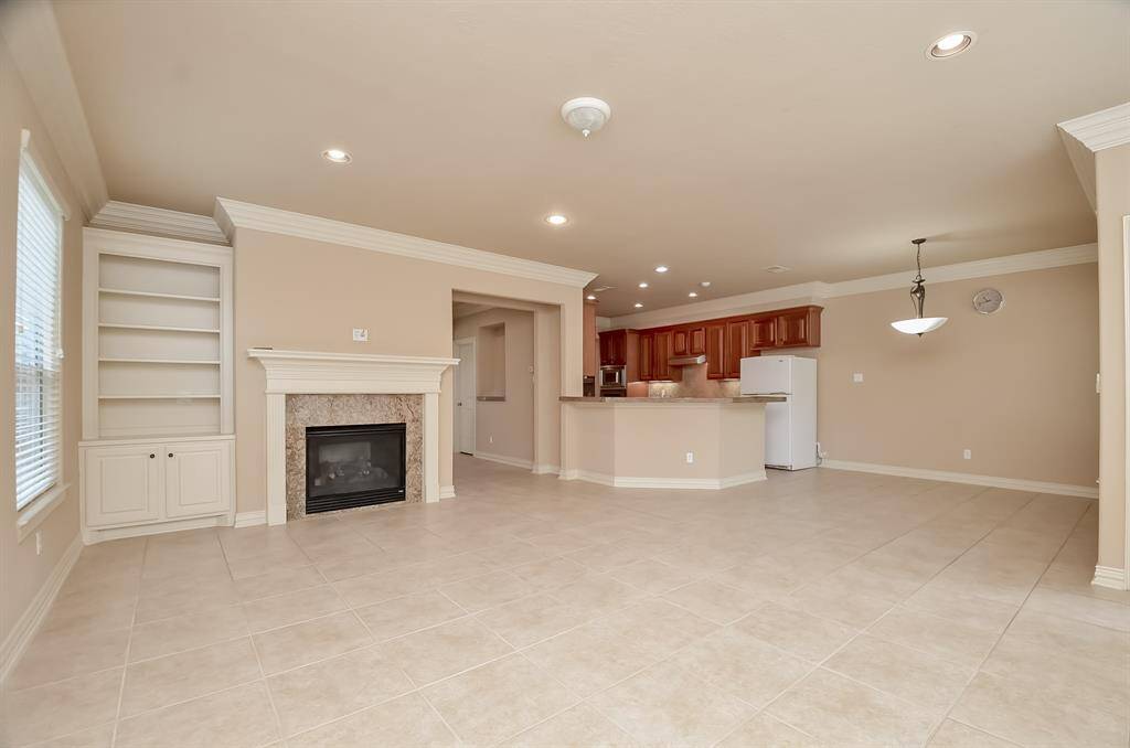 This spacious, open-concept living area features neutral tile flooring, a cozy fireplace with built-in shelving, and a modern kitchen with wooden cabinetry and recessed lighting. Perfect for entertaining and family gatherings.