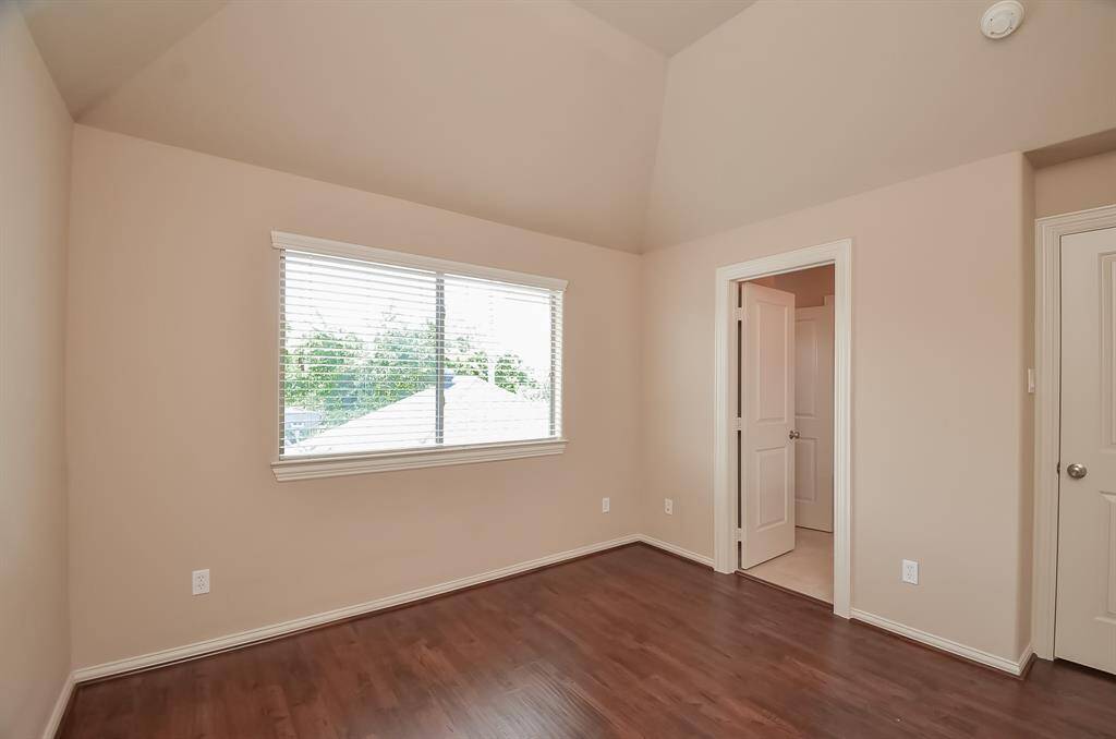 This room features warm wood flooring, a large window with blinds for natural light, and neutral wall colors. It has a high ceiling and includes a doorway leading to an adjacent area, ideal for use as a bedroom or office.