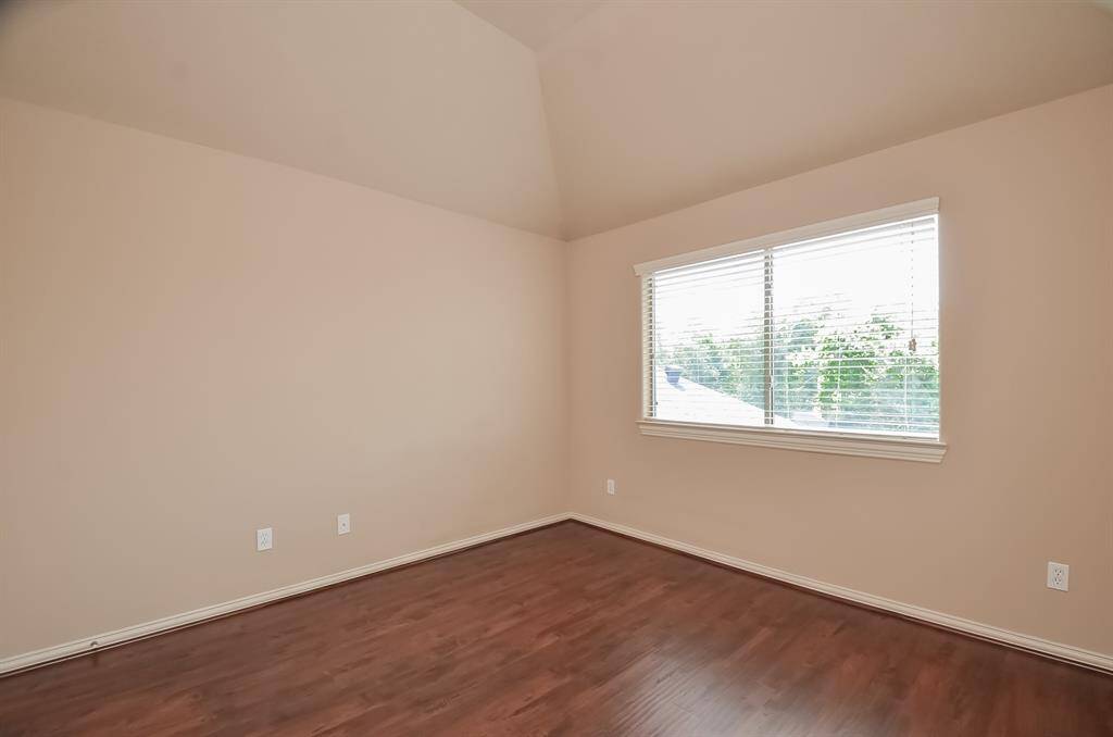 This room features neutral-colored walls, a vaulted ceiling, and rich hardwood flooring. It has a large window with blinds, allowing plenty of natural light. Perfect for a bedroom or home office.