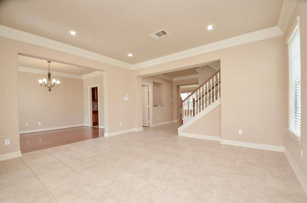 This spacious living area features neutral tile flooring, recessed lighting, and a light-filled open layout. It connects to a dining room with a chandelier and a staircase, providing a welcoming and versatile space for any home buyer.