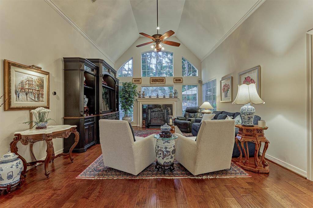 This elegant family room features high vaulted ceilings, a cozy fireplace, and large windows that bring in natural light. It is tastefully appointed with wood flooring, ceiling fan, and crown molding, and has a warm, inviting atmosphere.