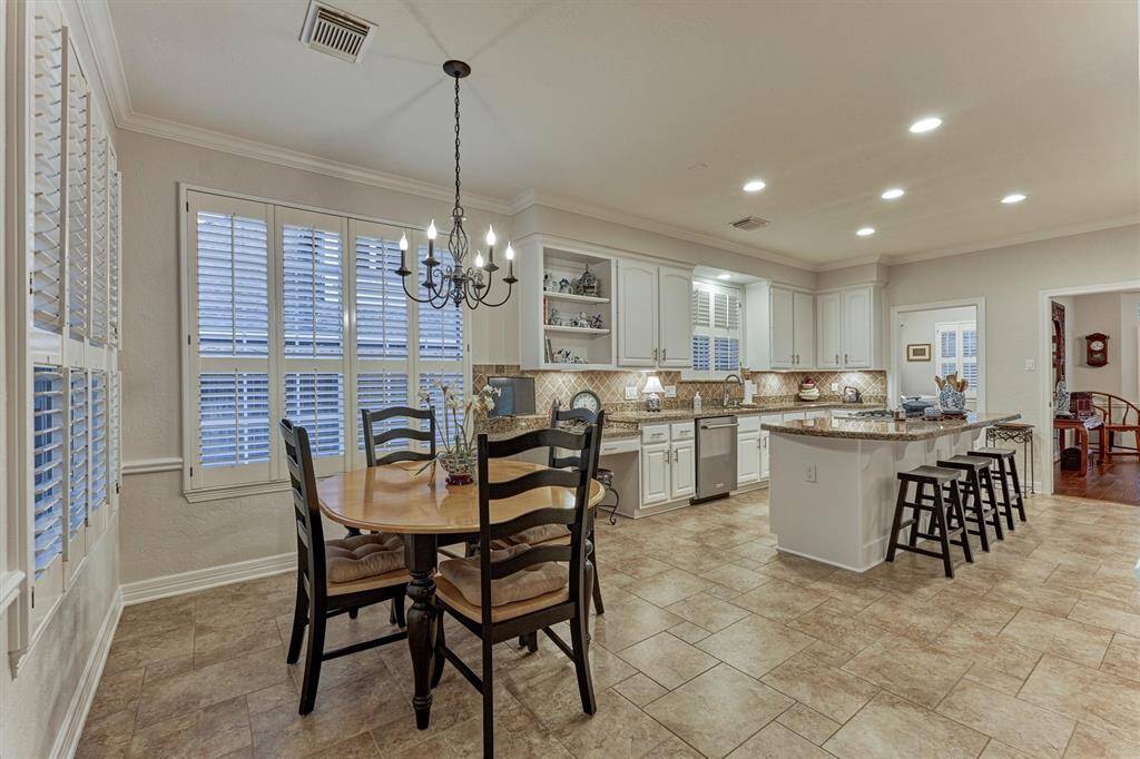 This spacious kitchen area features white cabinetry, granite countertops, and a large island with seating. It includes a cozy breakfast area with a chandelier and ample natural light from the windows with shutters. The open layout is perfect for entertaining and family gatherings.