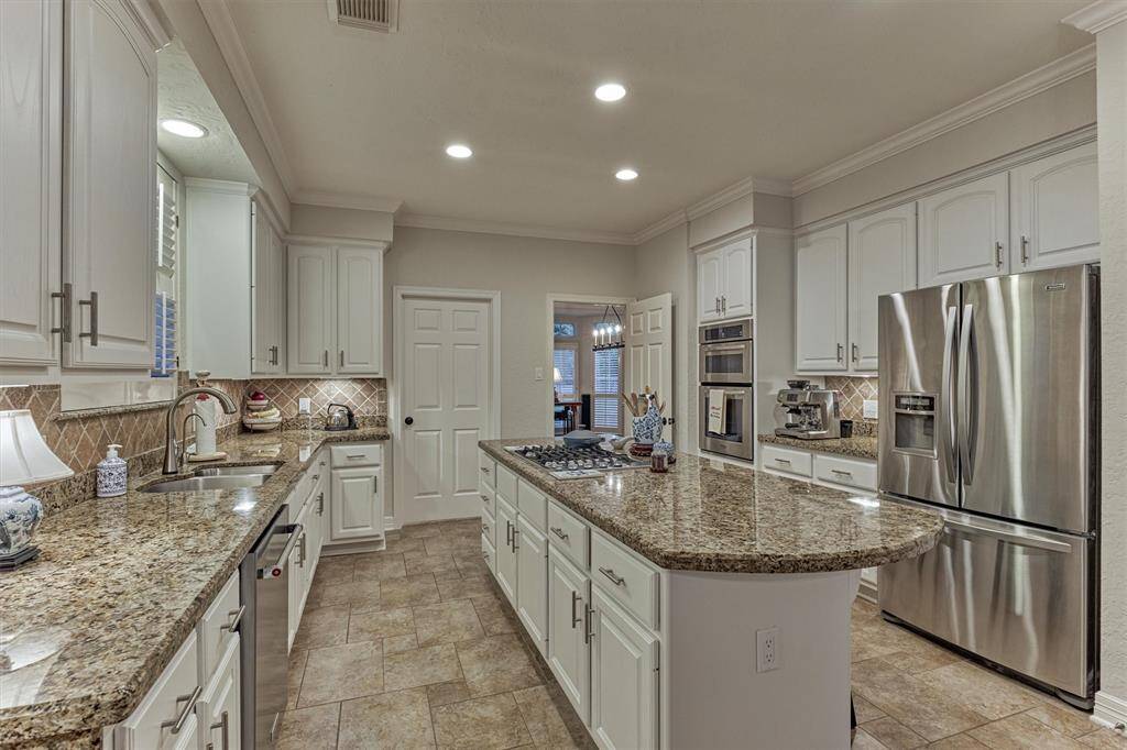 This kitchen features granite countertops, stainless steel appliances, and ample white cabinetry. The space includes a central island with a built-in cooktop and recessed lighting, offering a modern and functional layout.