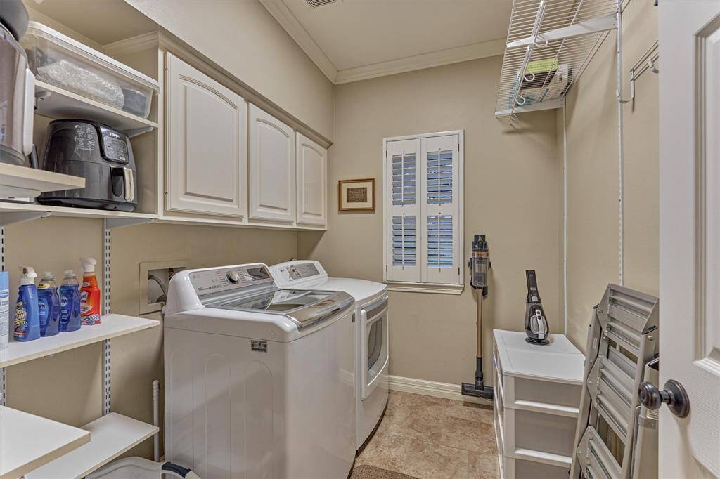 This photo shows a well-organized laundry room with a washer and dryer, ample cabinetry for storage, and additional shelving. The room is bright and functional, featuring a window with shutters for natural light, and tiled flooring for easy maintenance.