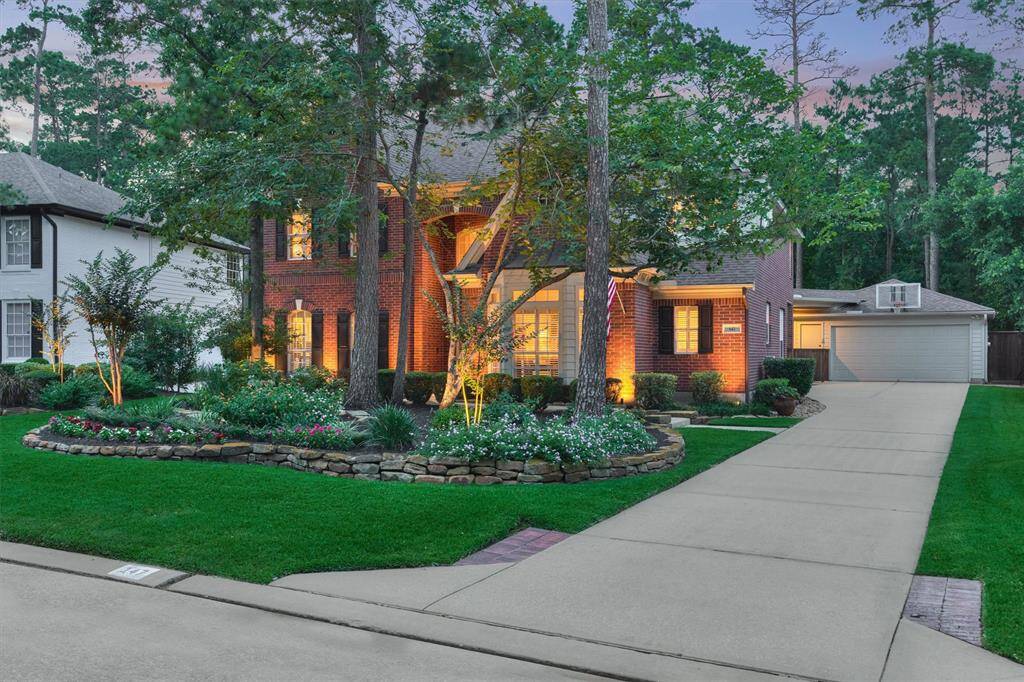 Prestine two-story brick home with a well-manicured lawn and mature trees. Features a spacious driveway leading to a detached garage, beautifully landscaped garden beds, and warm exterior lighting for an inviting ambiance.