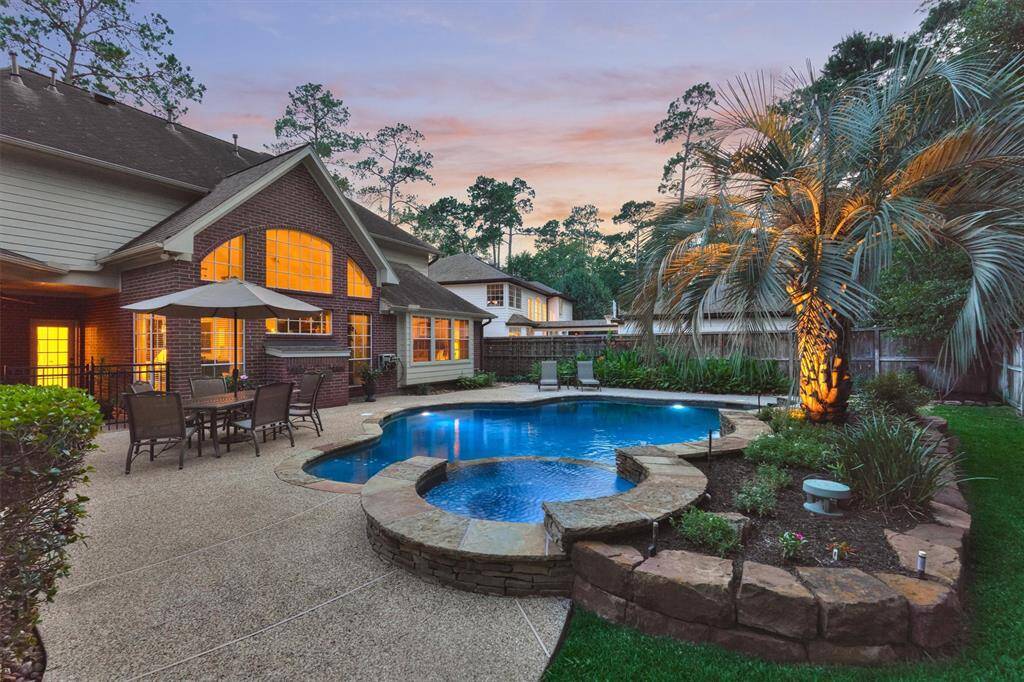 This photo showcases the beautiful backyard and well-lit pool and spa with extensive decking around the pool. The lush landscaping and surrounding greenery, creates a serene and inviting atmosphere.