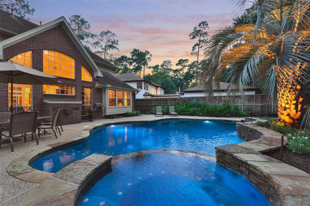 This stunning home features a beautifully lit backyard with a luxurious pool surrounded by lush landscaping. Large windows provide a warm view into the house, perfect for relaxation and entertaining.