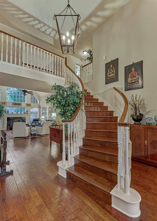 This inviting foyer features a grand staircase with white balusters and a stylish hanging light fixture. The space opens to a cozy living area with high ceilings, large windows, and a fireplace, offering a warm and elegant welcome.