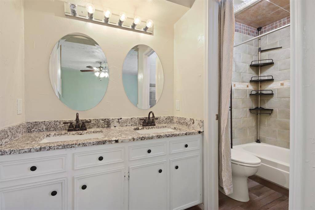 This incredible Primary Bathroom features wood tile floors, white cabinets, granite countertops, vanity lights, and his/her sinks with large round mirrors and sleek features.