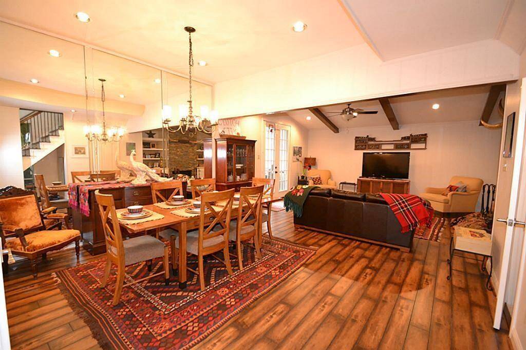 This inviting open-concept living and dining area features warm wood flooring, exposed beams, and ample natural light. The space is tastefully decorated with elegant chandeliers, a large dining table, and comfortable seating, perfect for entertaining or relaxing.