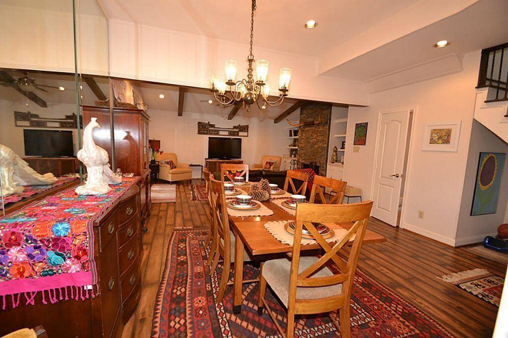This inviting dining and living area features warm wood flooring, a large dining table, and a cozy seating space with a stone fireplace. The room is well-lit with a stylish chandelier and recessed lighting, and decorated with vibrant textiles and artwork.