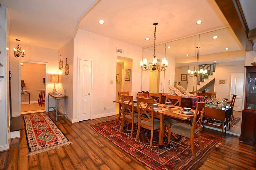 The photo showcases a spacious dining area with warm wood flooring and elegant chandeliers. It features a large dining table set for eight, surrounded by tasteful decor and mirrors that enhance the room’s brightness. The adjacent hallway and visible stairway add to the open feel, making it an inviting space for entertaining.