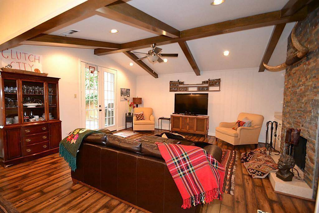 This cozy living room features a vaulted ceiling with wooden beams, a stone fireplace, and a mix of comfortable seating. Natural light pours in through French doors, leading to an inviting outdoor space. The room is accented with rustic decor and warm wood flooring, creating a welcoming atmosphere.