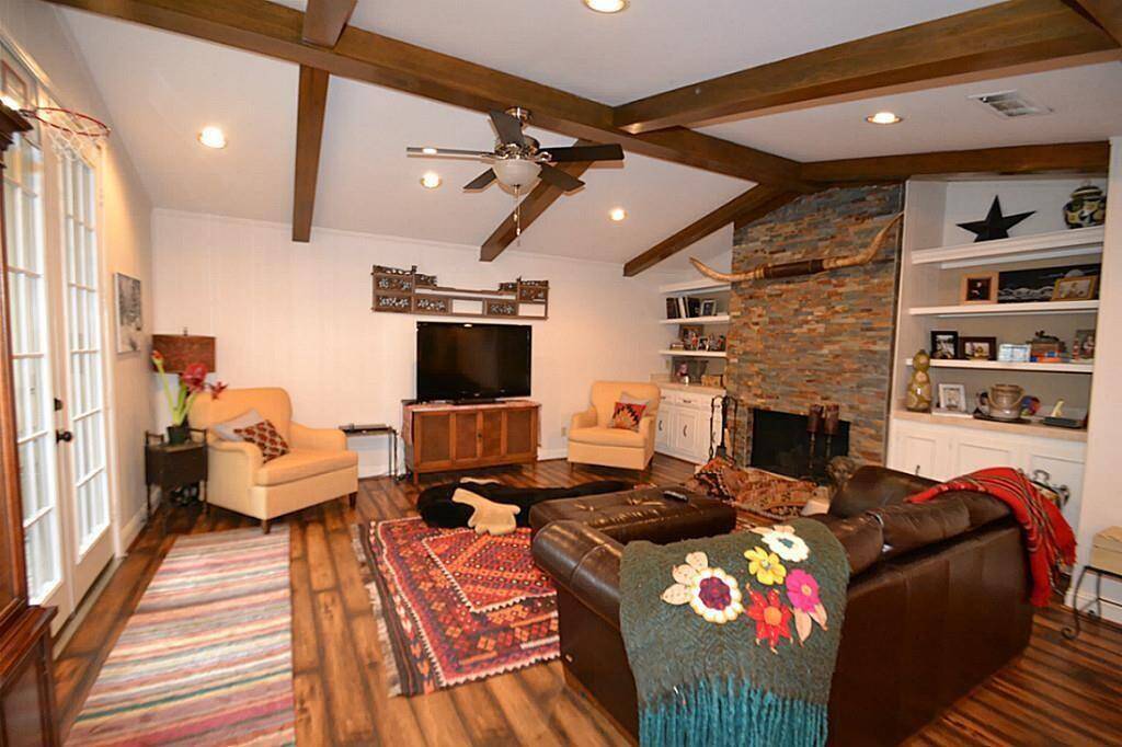 This cozy living room features a vaulted ceiling with wooden beams, a stone fireplace, and built-in shelving. It has a warm, inviting atmosphere with comfortable seating, hardwood floors, and French doors that let in natural light.