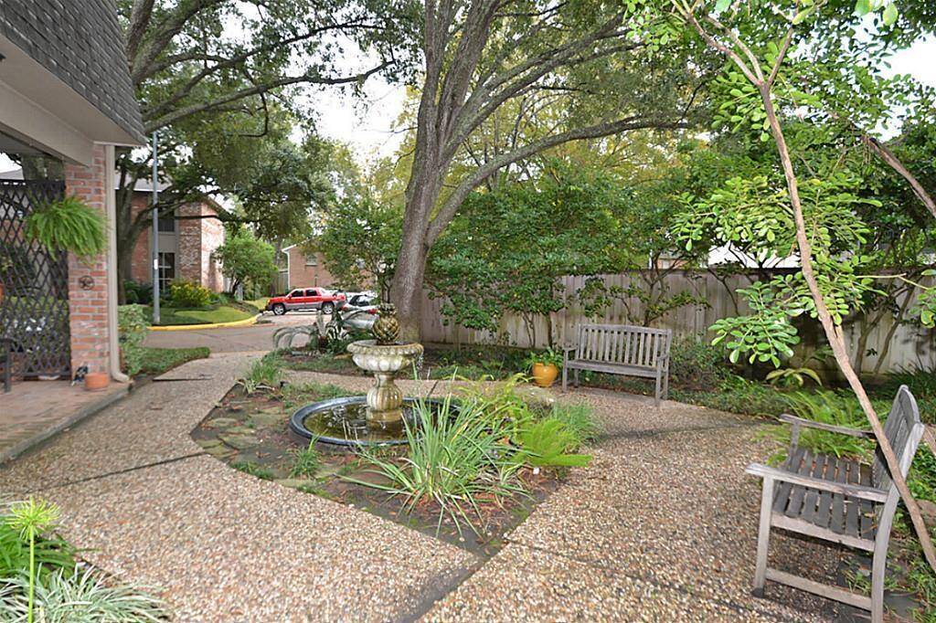 Charming garden space with a central fountain, surrounded by lush greenery and mature trees. Features seating areas for relaxation on a pebbled pathway, offering a serene outdoor retreat.