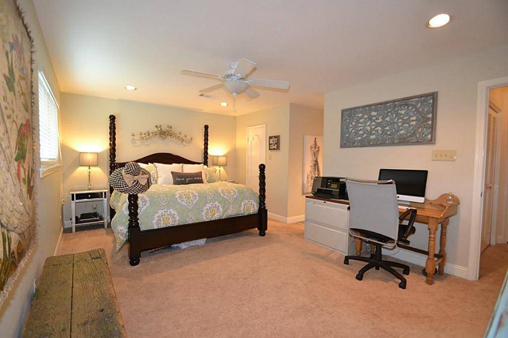 This spacious bedroom features a cozy bed with elegant posts, soft carpeting, and warm lighting. It includes a functional workspace with a desk and chair, and ample natural light from a window on the left. The decor is modern and inviting.