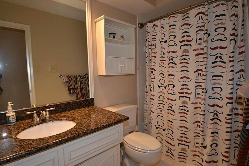 This bathroom features a granite countertop with a sink and a modern faucet. It includes a toilet, wall-mounted storage, and a shower with a playful mustache-themed curtain. The room is well-lit and has a neutral color scheme.
