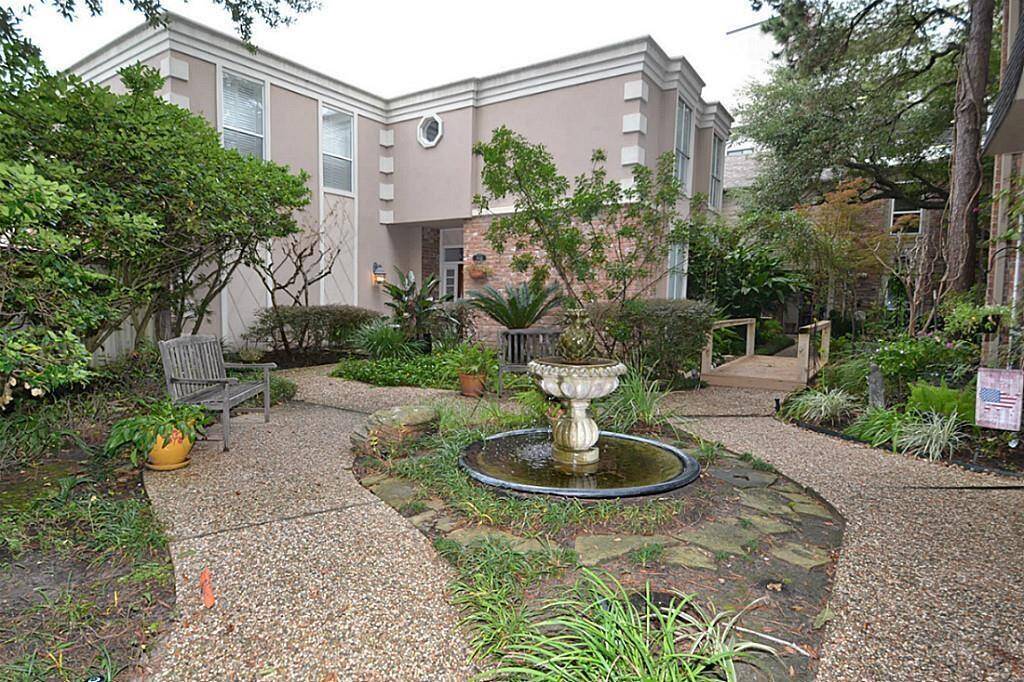 This charming courtyard features a well-maintained garden with lush greenery and a central fountain. The pathway is paved with pebbles, leading to a modern two-story home with large windows, offering a serene and inviting atmosphere.