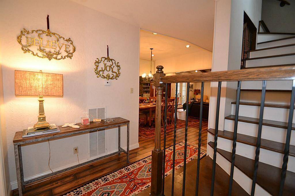 This welcoming entryway features warm wood flooring, a stylish table with a decorative lamp, and a staircase with elegant railings. The hallway leads to a dining area with ample natural light and cozy décor.