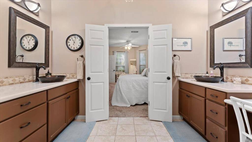 The guest en-suite bathroom features dual vanities, each featuring stylish vessel sinks and framed mirrors. The room has warm wood cabinetry and a neutral tile floor, leading into a guest suite.
