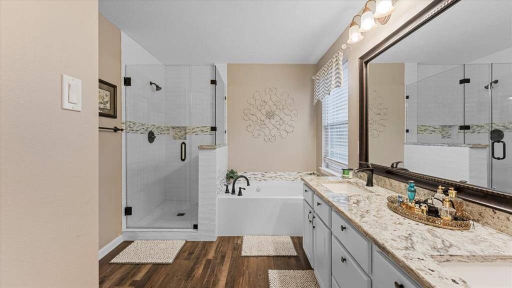 This primary bathroom features a spacious layout with a seamless shower and a jetted bathtub. It has a dual-sink granite countertop, elegant lighting, and luxury vinyl flooring, creating a modern and inviting space.