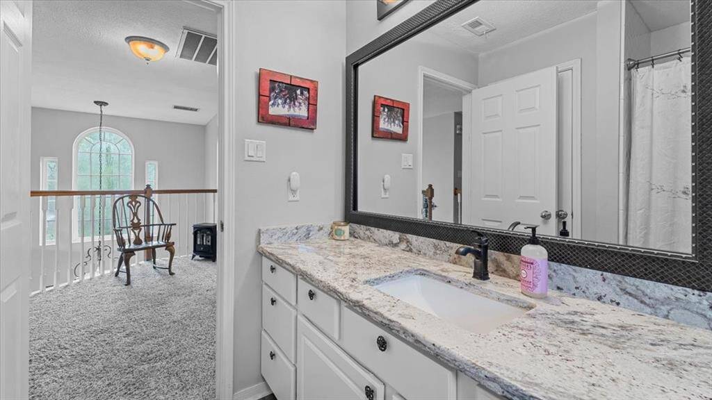 This upstairs bathroom offers a large granite countertop, plenty of drawers, and a sleek framed mirror. Adjacent is a cozy carpeted landing area, illuminated by natural light from an arched window.