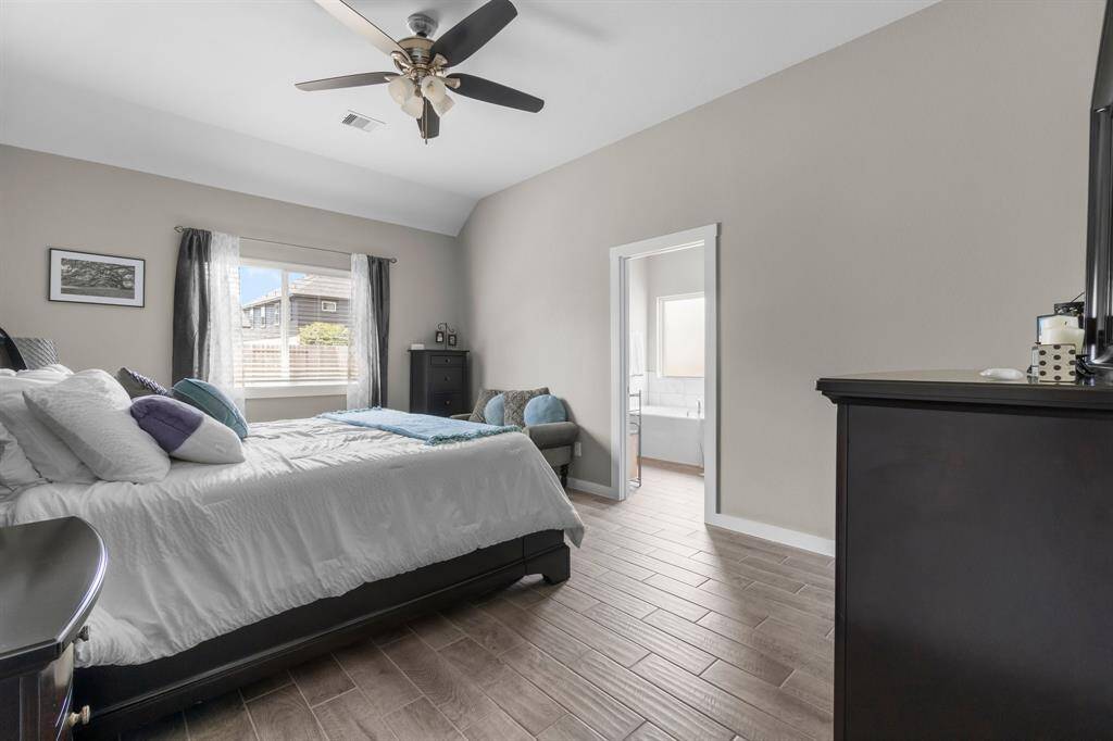 This bedroom features a neutral color palette with wood-look tile flooring, a ceiling fan, and a large window for natural light. It offers an ensuite bathroom for convenience and privacy.