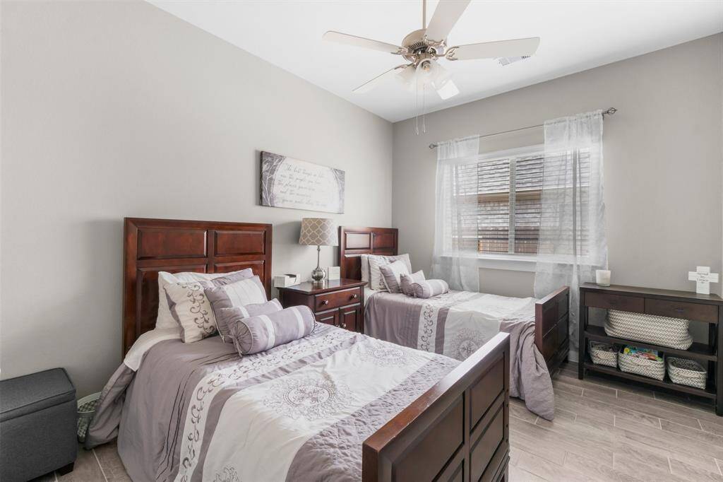 This cozy bedroom features two twin beds with matching wooden headboards and neutral bedding. A ceiling fan, soft lighting, and a window with sheer curtains create a bright, airy atmosphere. Ideal for guests or children with a large walking closet.