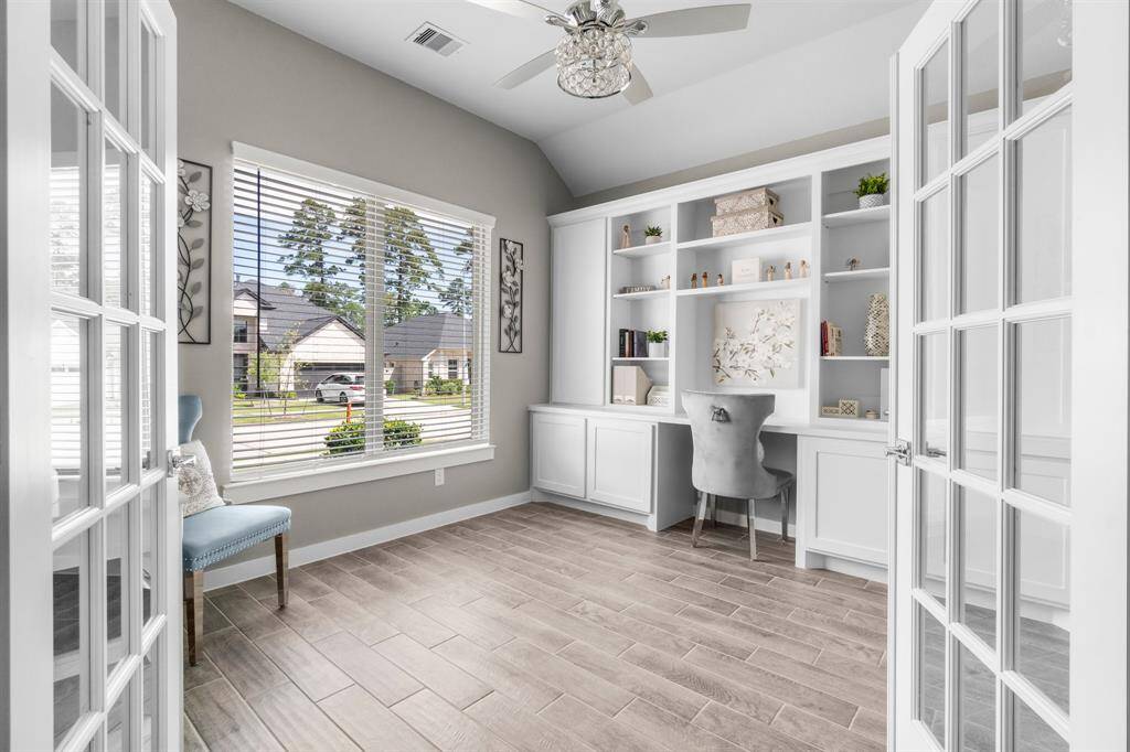 This home office features elegant French doors, a large window providing natural light, built-in white shelving, and a cozy desk area, creating a bright and inviting workspace.