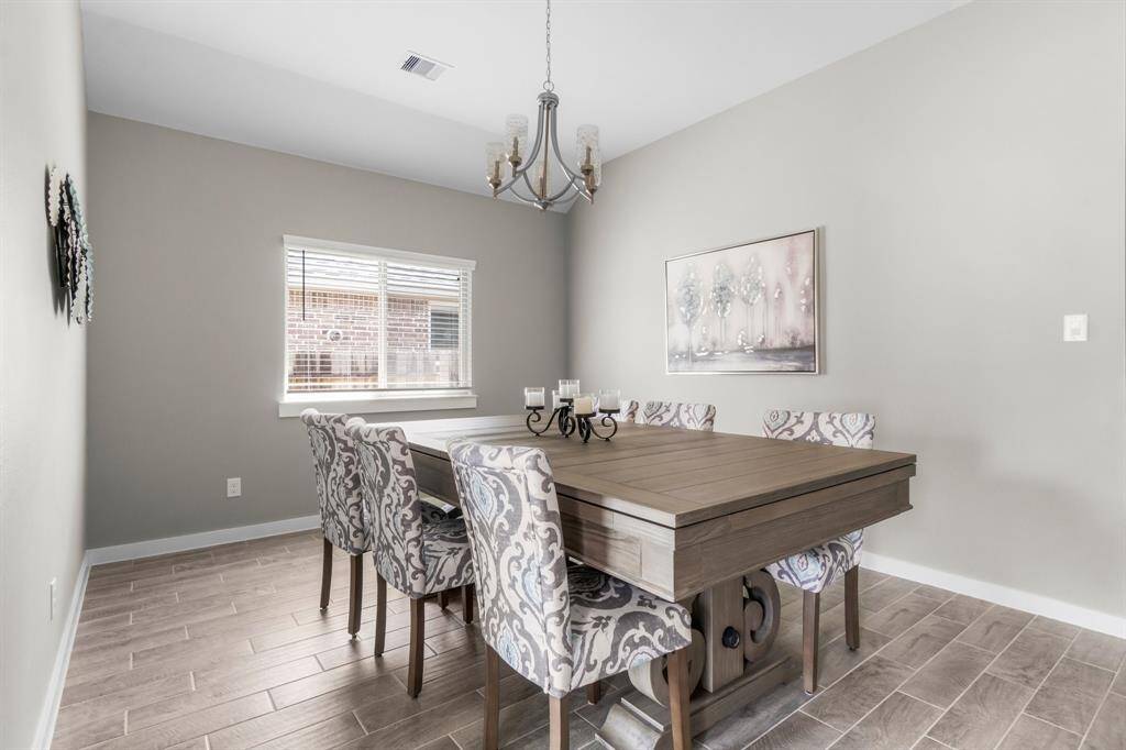 This is a bright dining room featuring a large wooden table with elegant patterned chairs. It has neutral walls, a modern chandelier, and a window that brings in natural light. This space has a clean, welcoming atmosphere.