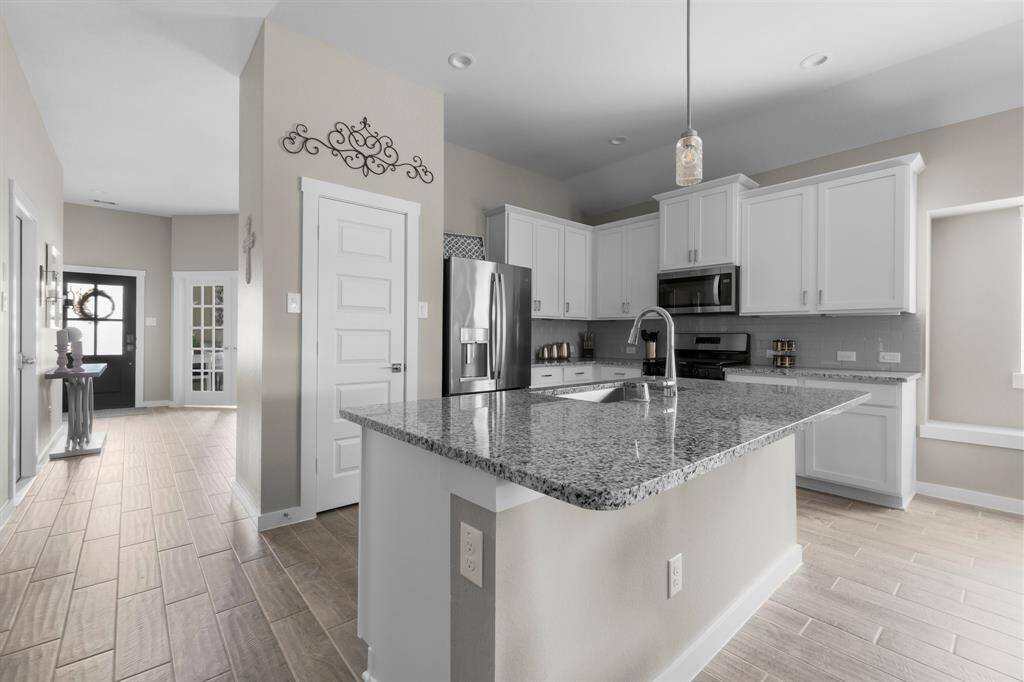 This photo showcases a modern kitchen with white cabinetry, a granite countertop island, and stainless steel appliances. The open layout leads to a bright hallway with wood-look tile flooring, creating a welcoming and functional space.