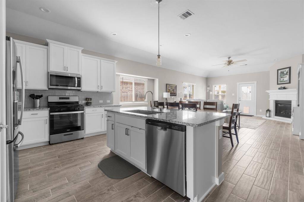 This spacious open-concept kitchen and living area features modern appliances, a large island with a sink, wood-look tile flooring, and ample natural light. It seamlessly connects to a cozy living room with a fireplace, creating an inviting atmosphere for entertaining or family gatherings.