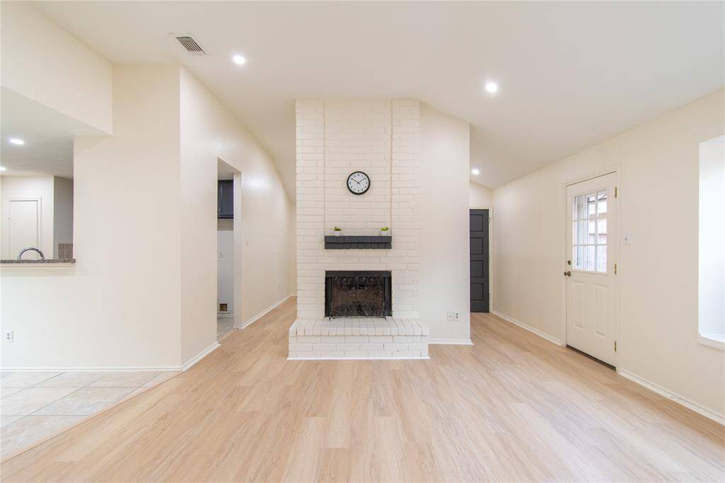 This bright living area features a cozy white brick fireplace as a focal point, complemented by light wood flooring and neutral walls. The space offers recessed lighting, a partially open floor plan with sightlines to the kitchen, and convenient access to the outdoors through a side door.