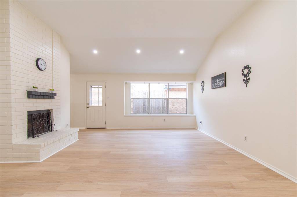 Living room with light wood flooring and a cozy brick fireplace. Recessed lighting and a large window enhance the airy atmosphere. Minimalist decor with wall accents and a welcoming vibe.