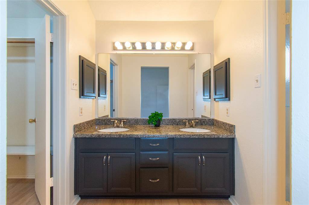 Modern bathroom with a double sink vanity featuring granite countertops and dark cabinetry. A large mirror is flanked by mounted lights, and there's a spacious closet visible to the left.