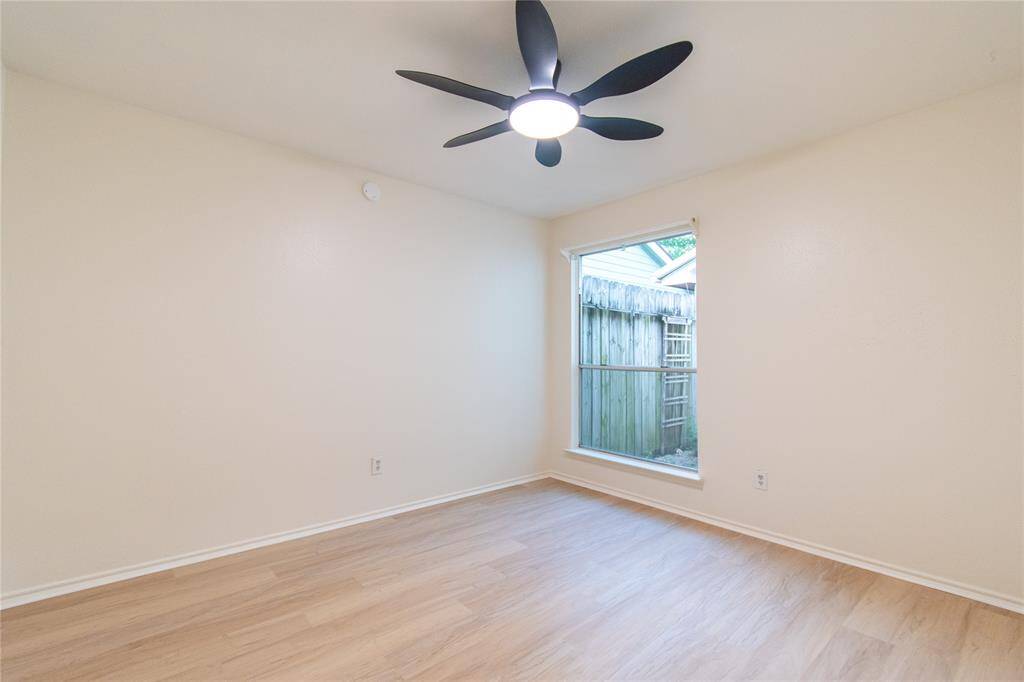 Bedroom 2  features light wood flooring, neutral walls, a modern ceiling fan, and a large window providing natural light and a view of a fenced outdoor area.