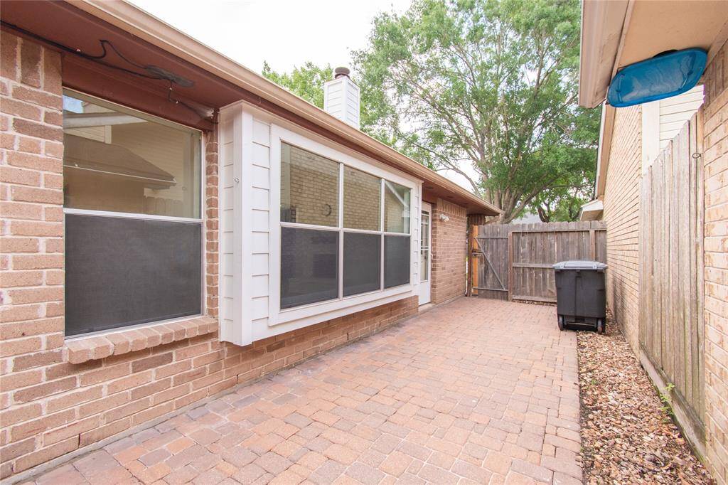 Low-maintenance brick patio area with large windows, providing natural light. It's enclosed by a wooden fence, offering privacy, and features neat landscaping with a paved walkway.