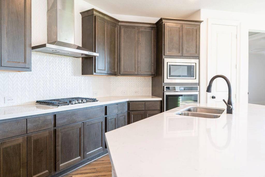 This modern kitchen features dark wood cabinetry, a sleek gas cooktop, built-in microwave and oven, and a stylish backsplash. It also includes a large island with a sink and elegant faucet, providing ample counter space for cooking and entertaining.