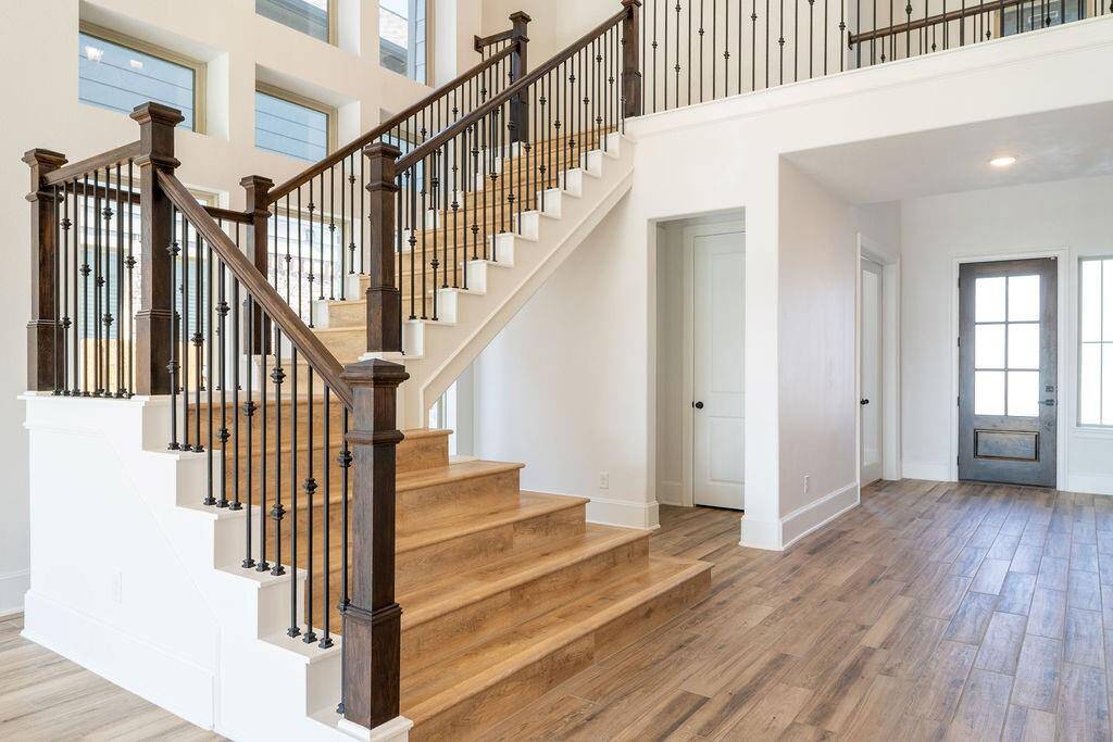 Modern, open foyer with elegant wooden stairs featuring wrought iron railings. The space is bright, with large windows and light wood flooring, creating an inviting entrance.