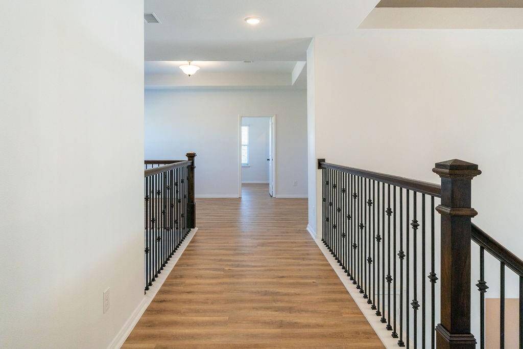 Well-lit, elevated hallway with wooden flooring and elegant black iron railings. The space opens into a room with a window at the end, creating an inviting and spacious feel connecting to the upstairs bedrooms