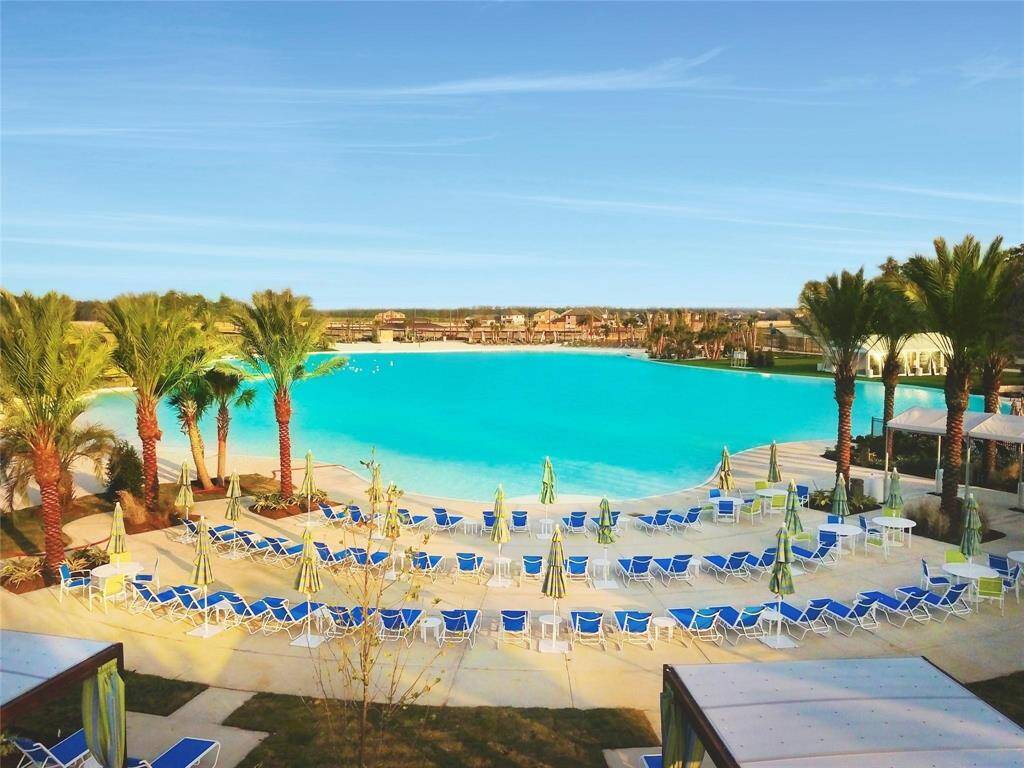 Walking distance to the resort-style pool area with a large, sparkling blue lagoon surrounded by palm trees. There are numerous lounge chairs and umbrellas for relaxation, offering a luxurious and inviting outdoor space.