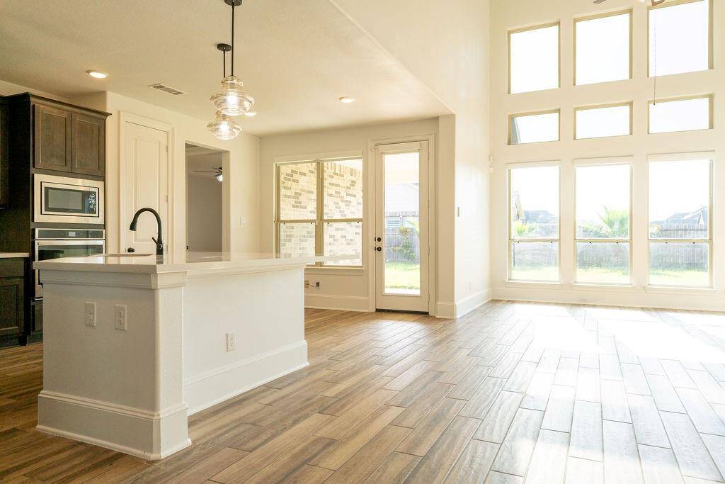 This bright and spacious living area features large windows, allowing abundant natural light. The open layout includes a modern kitchen with dark cabinetry, stainless steel appliances, and a sleek island, complemented by wood-look flooring throughout.