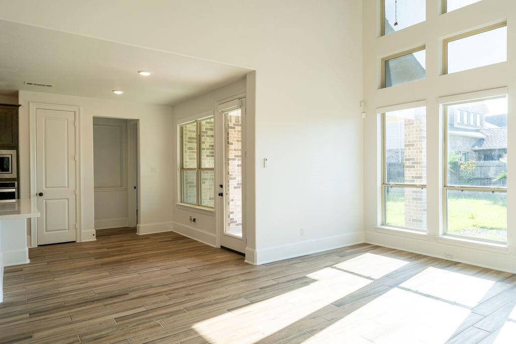 Bright and spacious room with large windows offering plenty of natural light. Features wood-like flooring, high ceilings, and an open layout leading to a kitchen area. Perfect for comfortable living and entertaining.