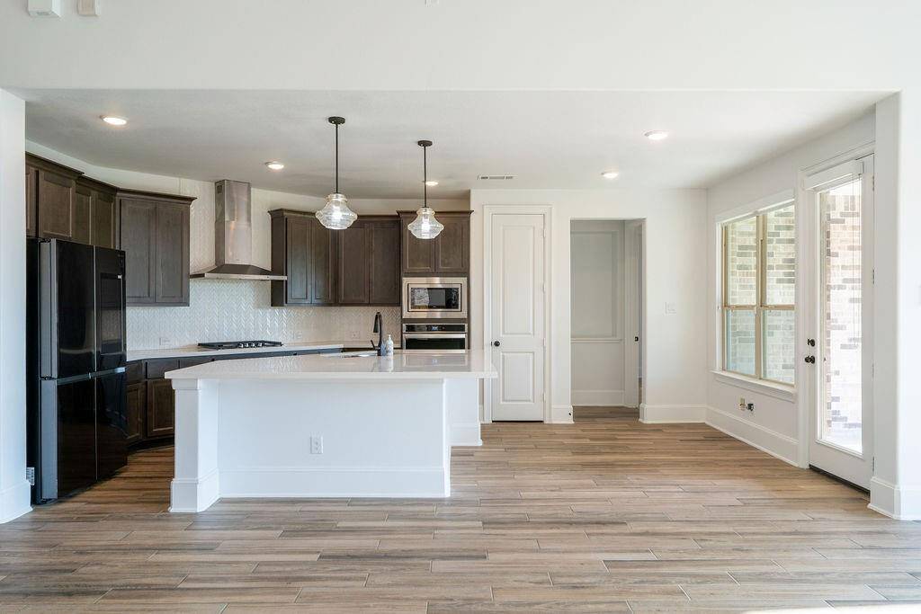 This modern kitchen features dark cabinetry, a sleek island with a white countertop, pendant lighting, and stainless steel appliances. The space is brightened by large windows and has wood-look tile flooring, creating a warm and inviting atmosphere.